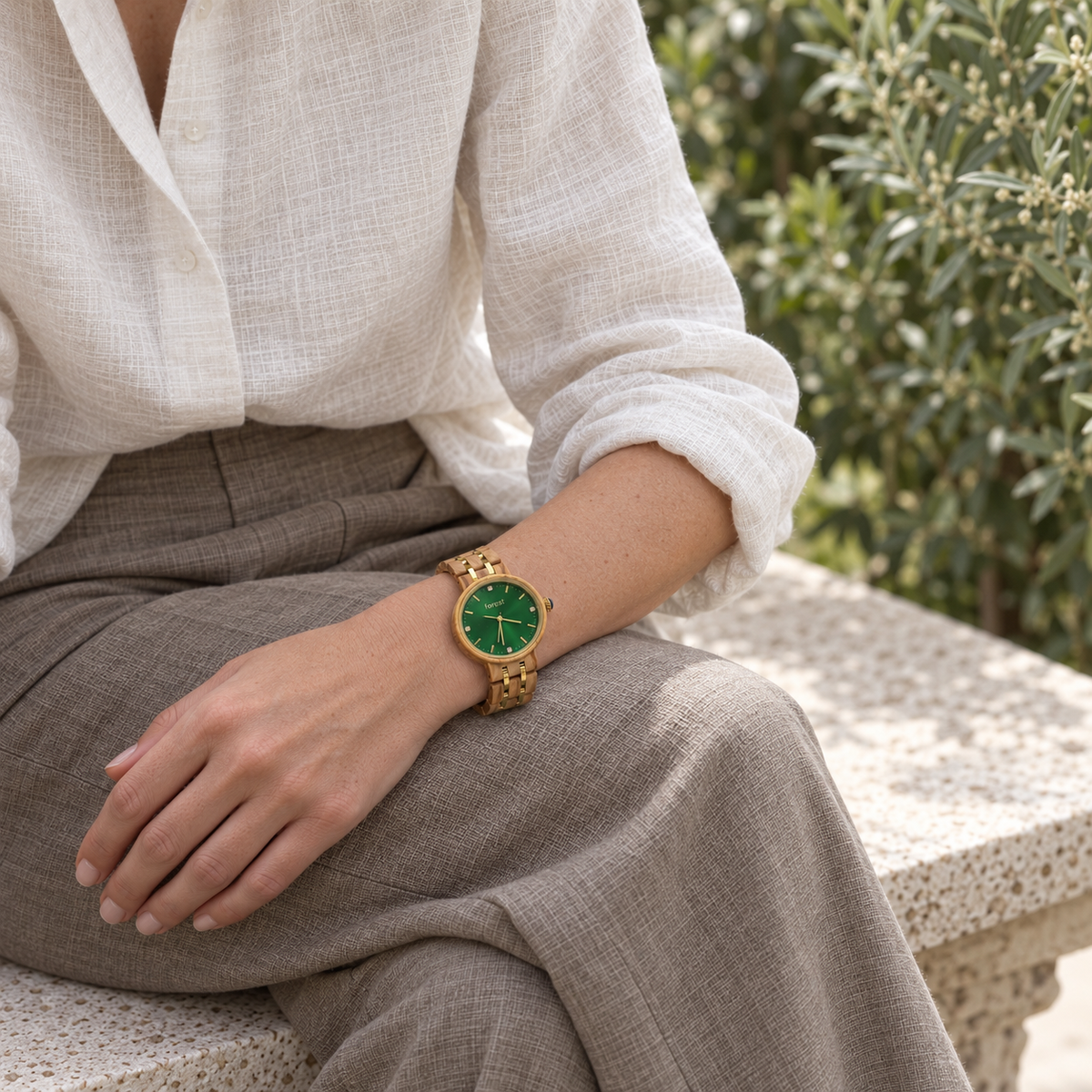 Ivy olivewood and gold-tone watch worn with an ivory linen shirt, seated on a stone bench in a quiet garden